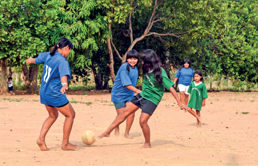 Imagem: Fotografia. Meninas, com blusas verdes e azuis chutando uma bola em um campo de terra. No fundo, há árvores. Fim da imagem.