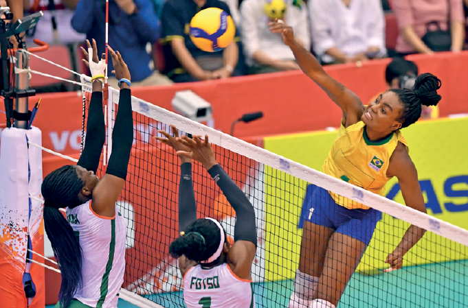 Imagem: Fotografia. Uma mulher com uniforme amarelo e azul, com o braço esquerdo estendido na direção de uma bola sobre uma rede. Do outro lado da rede, duas mulheres usando uniforme branco com os braços estendidos na direção da bola. Fim da imagem.