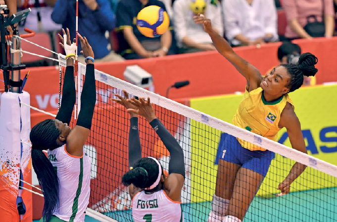 Imagem: Fotografia. Uma mulher com uniforme amarelo e azul, com o braço esquerdo estendido na direção de uma bola sobre uma rede. Do outro lado da rede, duas mulheres usando uniforme branco com os braços estendidos na direção da bola.  Fim da imagem.