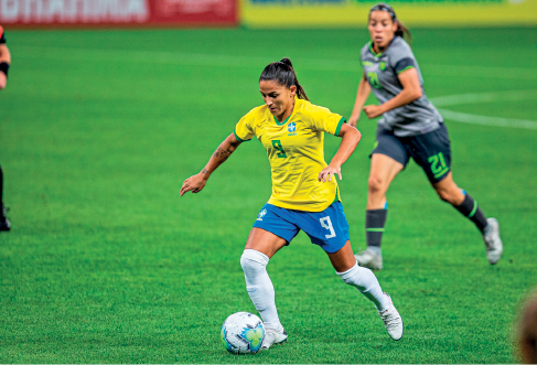 Imagem: Fotografia. Uma mulher com uniforme do Brasil, correndo com uma bola no pé em um campo com grama verde, atrás, há outra mulher com uniforme do Equador correndo.  Fim da imagem.