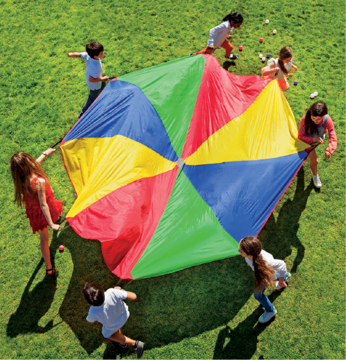 Imagem: Fotografia. Vista aérea. Sete crianças segurando um paraquedas colorido com as mãos. Elas estão de pé em um gramado caminhando em círculo.  Fim da imagem.