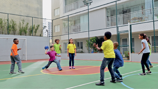 Imagem: Fotografia. Crianças em uma quadra de com prédios em volta. Um deles segura uma bola na direção das outras crianças.  Fim da imagem.