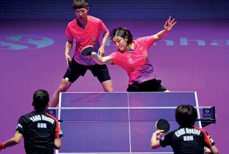 Imagem: Fotografia. Dois jogadores de costas, segurando raquete pequena, seguida de uma mesa com uma rede no meio. No fundo, do outro lado da mesa, dois jogadores com uniforme rosa, segurando raquetes.  Fim da imagem.