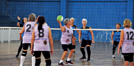 Imagem: Fotografia. Mulheres com cabelos brancos e rugas usando uniformes em uma quadra com rede baixa. Uma delas segura uma bola próxima à rede na direção do time adversário. Fim da imagem.