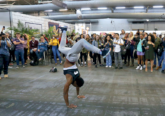 Imagem: Fotografia. 4. Passo com apoio dos dois braços. Nova York, Estados Unidos, 2019. Um homem usando regata e calça jeans. Ele está com as duas mãos apoiando o chão com as pernas flexionadas para cima com o tronco no ar. No fundo várias pessoas olhando para ele. Fim da imagem.