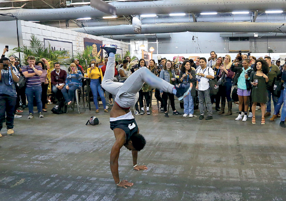 Imagem: Fotografia. 4. Passo com apoio dos dois braços. Nova York, Estados Unidos, 2019. Um homem usando regata e calça jeans. Ele está com as duas mãos apoiando o chão com as pernas flexionadas para cima com o tronco no ar. No fundo várias pessoas olhando para ele.  Fim da imagem.