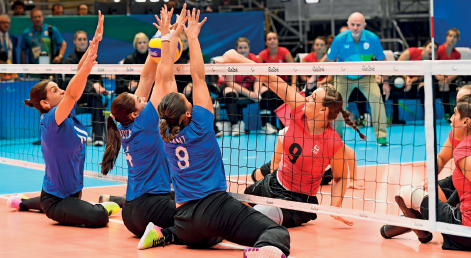 Imagem: Fotografia. À esquerda, três mulheres com uniforme azul, sentadas em uma quadra, com rede baixa. Elas estão com os braços estendidos na direção da bola próximo à rede. À direita, duas mulheres com uniforme laranja, uma delas está com o braço estendido na direção da bola, próximo à rede. No fundo, arquibancada com pessoas sentadas. Fim da imagem.