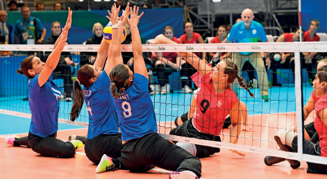 Imagem: Fotografia. À esquerda, três mulheres com uniforme azul, sentadas em uma quadra, com rede baixa. Elas estão com os braços estendidos na direção da bola próximo à rede. À direita, duas mulheres com uniforme laranja, uma delas está com o braço estendido na direção da bola, próximo à rede. No fundo, arquibancada com pessoas sentadas.  Fim da imagem.