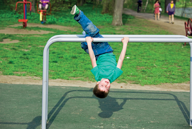 Imagem: Fotografia. Um menino usando camiseta verde e calça jeans. Ele está segurando uma barra com a cabeça para baixo e as pernas estendidas para cima.  Fim da imagem.