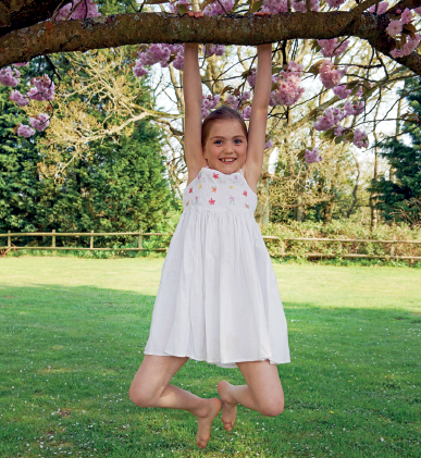 Imagem: Fotografia. Uma menina segurando com as duas mãos em uma árvore com os joelhos flexionados e os pés no ar. Fim da imagem.
