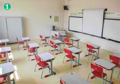 Imagem: Fotografia. Uma sala de aula. Na frente, mesa maior encostada na parede e uma lousa branca. Na parede do canto há um armário branco e na área da sala há carteiras com cadeiras vermelhas enfileiradas.  Fim da imagem.