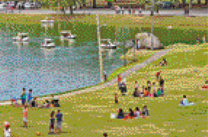 Imagem: Fotografia. Um parque gramado com pessoas sentadas em grupos. Ao lado há um lago com alguns barcos.    Fim da imagem.