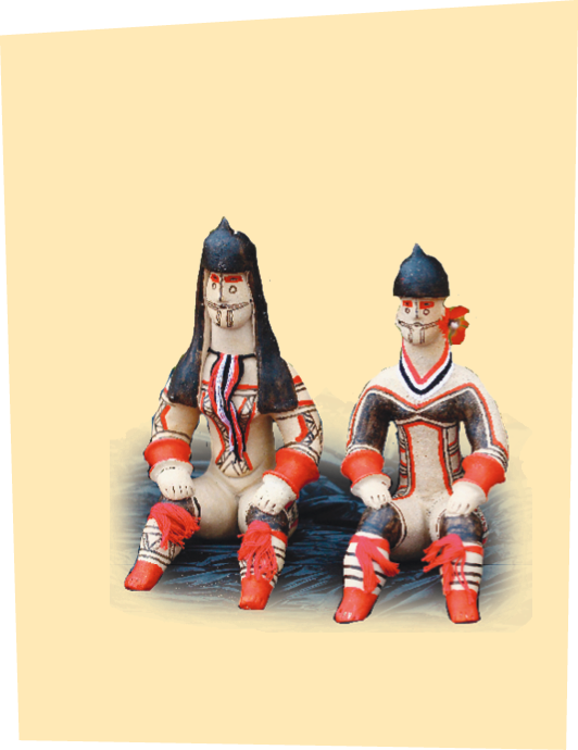 Imagem: Fotografia. Dois bonecos indígenas de cabelo preto e liso, com pinturas no rosto e no corpo. Eles estão sentados.   Fim da imagem.