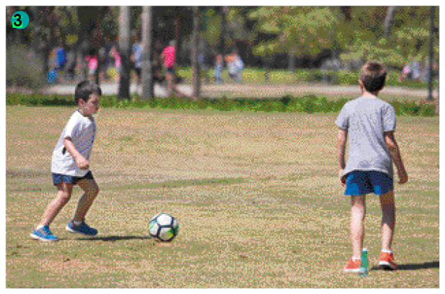 Imagem: Fotografia. Dois meninos jogando bola. Um tem o cabelo preto, bermuda azul e camiseta branca e o outro tem o cabelo castanho, bermuda vermelha e camiseta cinza.   Fim da imagem.
