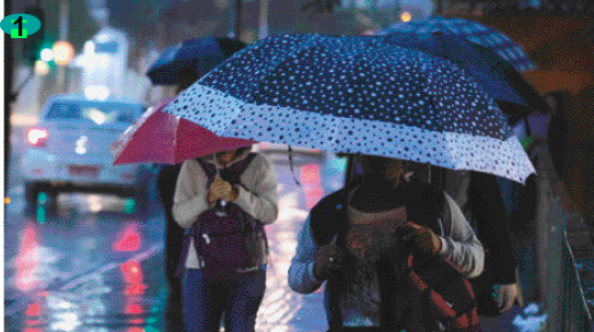 Imagem: Fotografia 1. Pessoas caminhando na chuva, em uma calçada, segurando guarda-chuva. É noite e ao lado delas tem a rua com carros.  Fim da imagem.