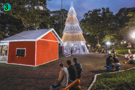 Imagem: Fotografia 3. Praça com uma pequena casa, uma árvore de Natal com luzes e pessoas sentadas em bancos. Fim da imagem.