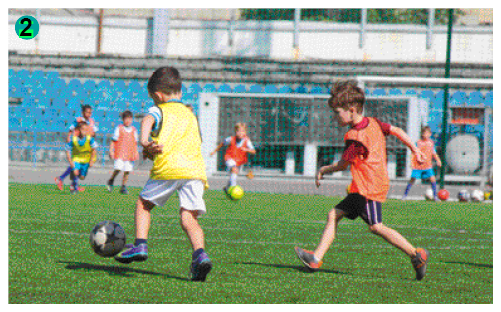 Imagem: Fotografia 2. Crianças jogando futebol em um campo gramado. Alguns meninos usam colete laranja e alguns usam colete amarelo.   Fim da imagem.