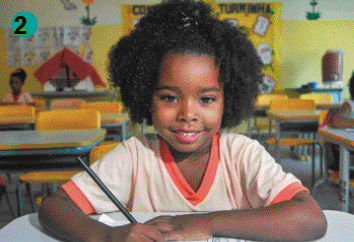 Imagem: Fotografia. Uma menina negra de cabelo preso no alto da cabeça vestindo uma camiseta laranja. Ela está sentada em uma carteira em uma sala de aula e escreve em um caderno.  Fim da imagem.