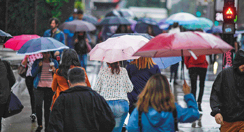 Imagem: Fotografia. Pessoas andando em uma calçada. Algumas delas seguram guarda-chuvas de diversas cores.  Fim da imagem.