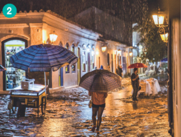 Imagem: Fotografia. Uma mulher de calça e casaco de costas segurando um guarda-chuva. Ao lado dela há um carrinho embaixo de um guarda-chuva e mais para frente um homem parado. Ela caminha em uma rua de pedras embaixo da chuva. Dos lados tem construções antigas brancas com janelas e portas em azul. Fim da imagem.