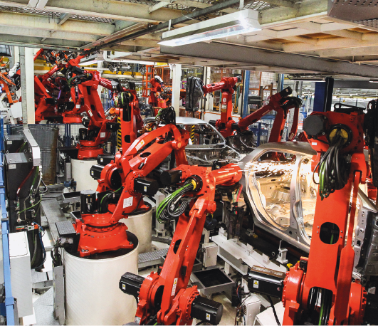 Imagem: Fotografia. Uma fábrica com robôs montadores laranjas.  Fim da imagem.
