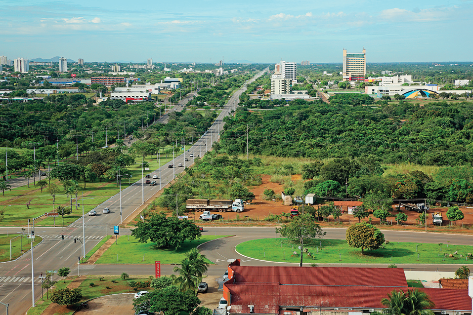 Imagem: Fotografia. Vista aérea de uma cidade com uma grande avenida principal, com fileiras de postes de iluminação do lado. Há grandes áreas cobertas por árvores e no fundo algumas casas e prédios.  Fim da imagem.