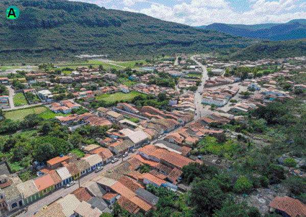 Imagem: Fotografia. Vista aérea de uma cidade, com prédios, casas e áreas com vegetação. Fim da imagem.