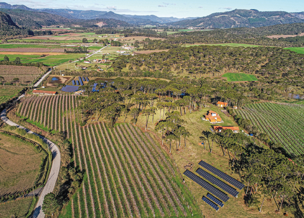 Imagem: Fotografia. Paisagem com plantações, arvores e painéis de energia solar. Para a direita tem algumas casas e no fundo da paisagem uma cadeira de montanhas.  Fim da imagem.