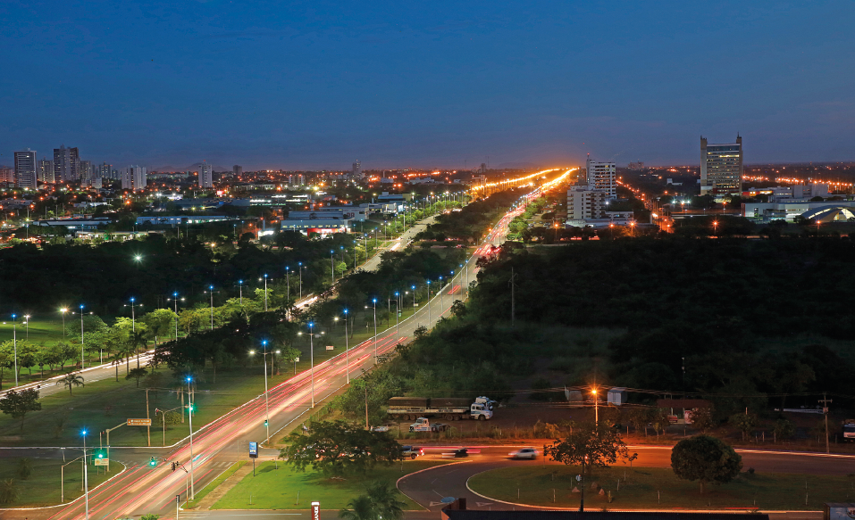 Imagem: Fotografia. Vista aérea de uma cidade durante a noite com uma grande avenida principal, com fileiras de postes de iluminação acesos do lado. Há grandes áreas cobertas por árvores e no fundo algumas casas e prédios.  Fim da imagem.