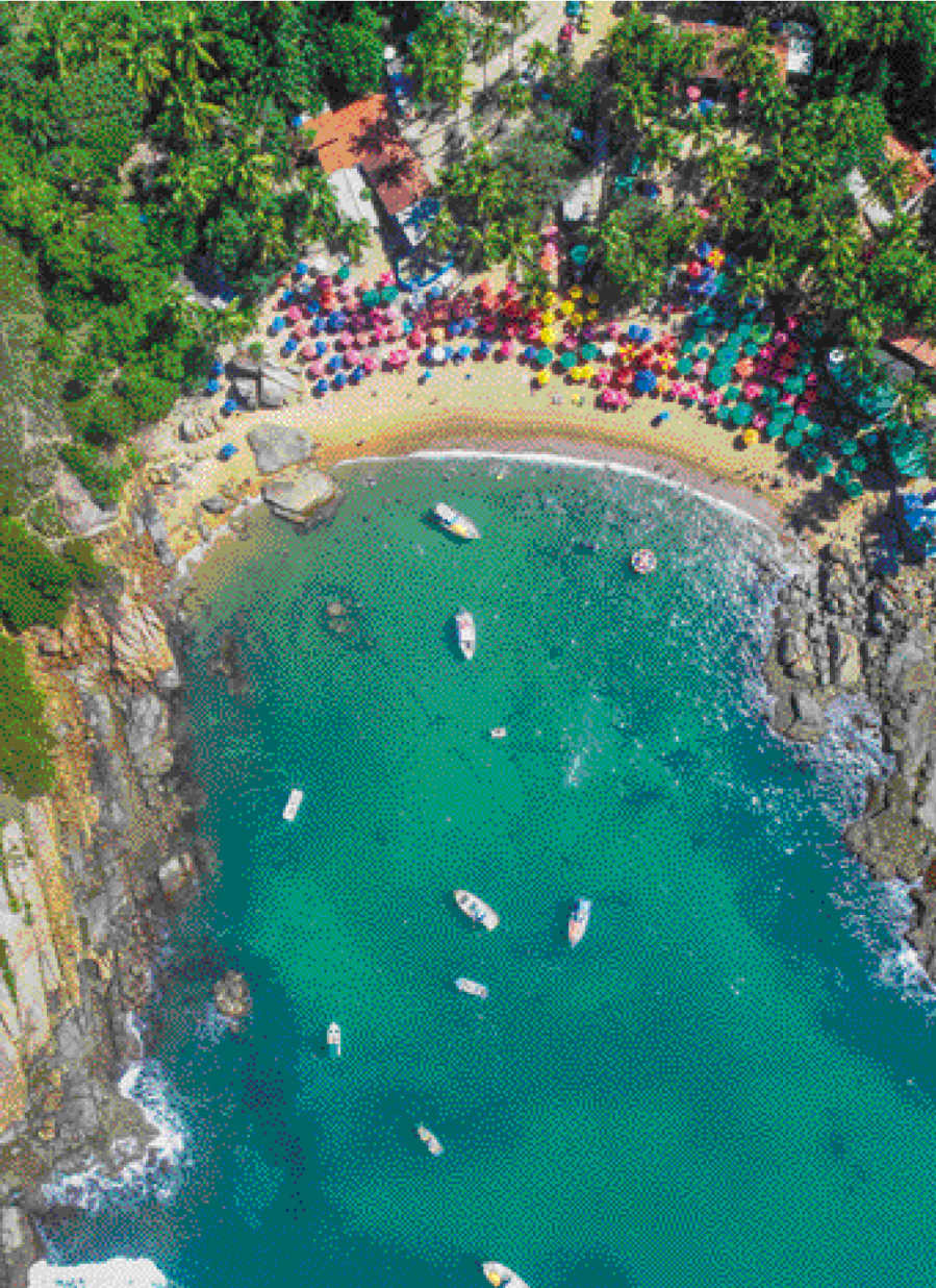 Imagem: Fotografia. Uma praia vista de cima. O mar é azul claro com alguns barcos e de cada um dos lados há pedras. A areia está coberta com pessoas e guarda-sóis coloridos. Atrás da praia tem uma área com árvores e algumas casas.  Fim da imagem.