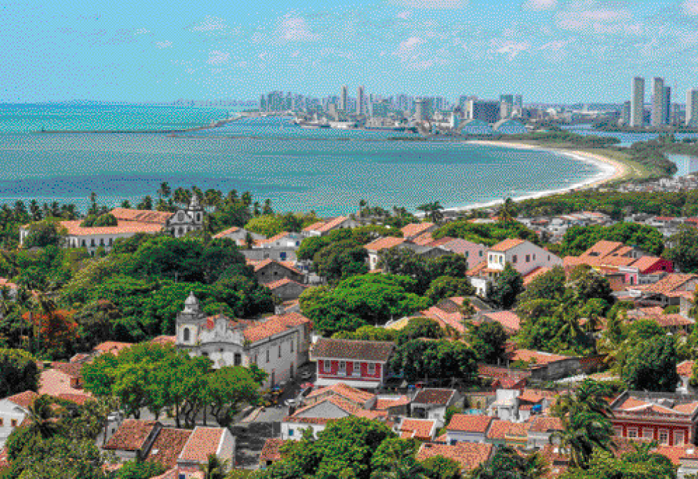 Imagem: Fotografia. Uma cidade vista de cima, com casas, uma igreja e muitas árvores entre elas. No fundo, o mar e uma faixa de areia e vegetação do lado direito que vai até uma segunda parte da cidade, cheia de prédio.  Fim da imagem.