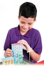 Imagem: Fotografia. Um menino de cabelo curto preto usando uma camiseta roxa. Ele sorri e está com as mãos em um prédio azul feito com caixa.  Fim da imagem.