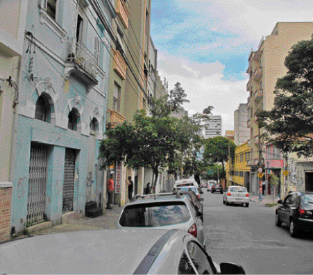 Imagem: Fotografia. Uma rua com carros estacionados e construções de diferentes cores. Nas calçadas há algumas pessoas e canteiros com árvores.  Fim da imagem.