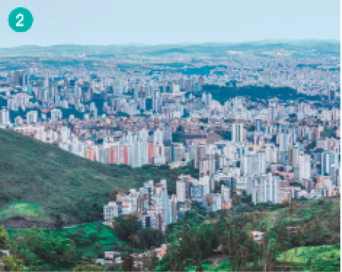 Imagem: Fotografia. Vista aérea de uma cidade cheia de prédios e com áreas gramadas em primeiro plano. Fim da imagem.