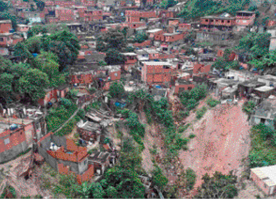 Imagem: Fotografia. Vista aérea de uma comunidade com as casas construídas ao redor de um deslizamento de terra.  Fim da imagem.