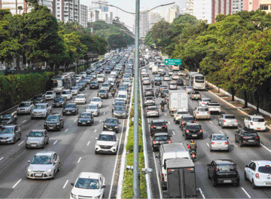 Imagem: Fotografia. Uma avenida com um canteiro no meio e trânsito nas duas direções. Nas margens da avenida há árvores.  Fim da imagem.