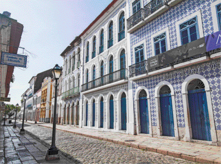 Imagem: Fotografia. Prédios com azulejos na fechada. A rua na frente deles é de pedra e tem postes de iluminação. Fim da imagem.