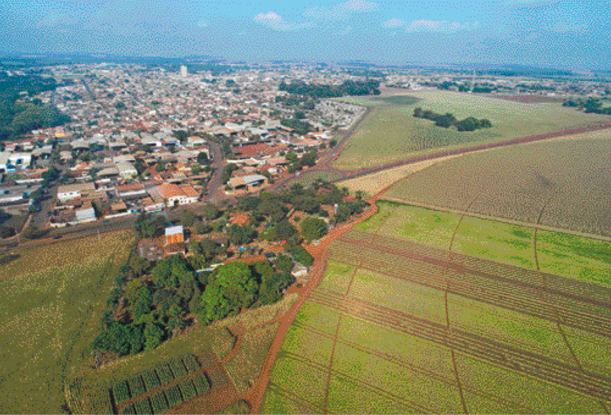 Imagem: Fotografia. Uma cidade com quadras com casas. Na frente e do lado direito, áreas de plantações.  Fim da imagem.