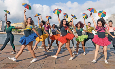 Imagem: Fotografia. Um grupo de pessoas na beira de um rio. Os meninos vestem calças e as meninas saias coloridas, eles seguram um guarda-chuva colorido em cima da cabeça.  Fim da imagem.