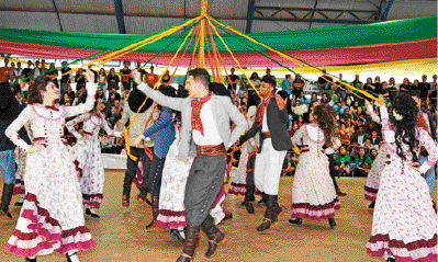 Imagem: Fotografia. Um grupo de pessoas dançando. Os homens usam calça, camisa e lenço vermelho no pescoço e as mulheres usam vestido longo. Eles seguram fitas colorias que estão presas a uma haste no meio. Fim da imagem.