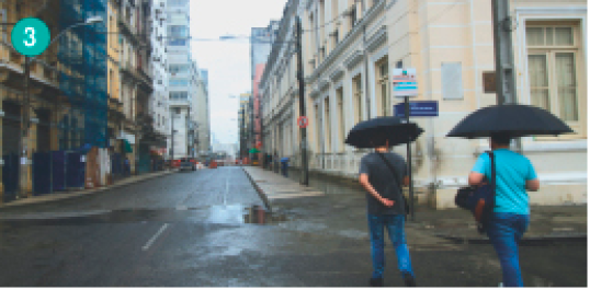 Imagem: Fotografia. Duas pessoas segurando um guarda-chuvas em uma calçada. A rua está vazia e dos lados tem prédios antigos.  Fim da imagem.