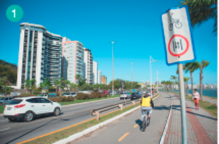 Imagem: Fotografia. Uma pessoa andando de bicicleta em uma ciclovia. A ciclovia está ao lado de uma rua com carros e do lado dela há prédios. Há uma placa com o desenho de uma bicicleta.  Fim da imagem.