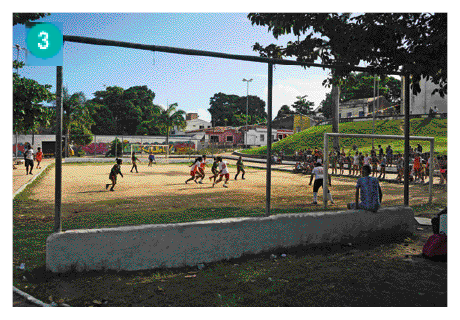 Imagem: Fotografia. Pessoas jogando futebol em um campo de areia. Do lado, pessoas assistem sentadas em uma arquibancada de madeira.  Fim da imagem.