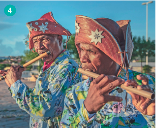 Imagem: Fotografia. Dois homens de cabelo branco tocando uma flauta de madeira. Ambos vestem uma camisa azul estampada e usam um chapéu marrom com estrelas.  Fim da imagem.