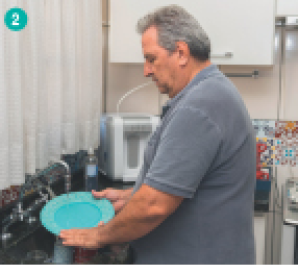 Imagem: Fotografia. Um homem de cabelo branco e camiseta cinza lavando louça. Fim da imagem.