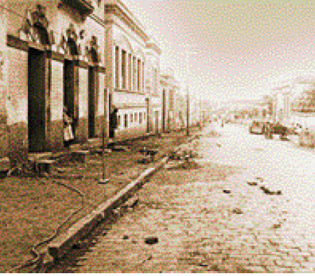 Imagem: Fotografia. Imagem em sépia de uma rua de pedra com as guias quebradas. A calçada está em obras, com carrinho de pedreiro. Do lado, construções antigas, sendo que na primeira delas tem uma mulher de vestido longo apoiada na porta.  Fim da imagem.