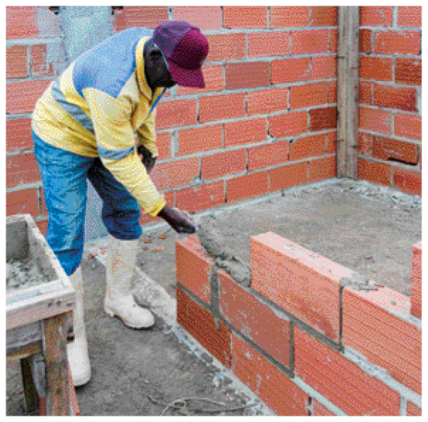Imagem: Fotografia. Um homem negro usando calça jeans, bota, uma camiseta e boné. Ele está passando cimento em uma parede de tijolos em construção.  Fim da imagem.