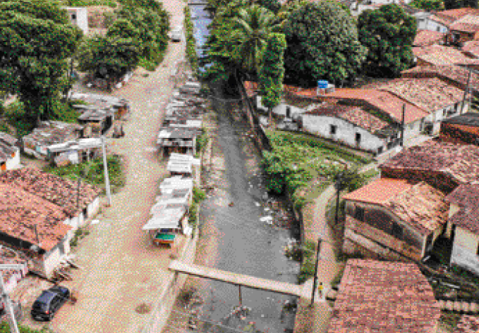 Imagem: Fotografia. Vista aérea de uma comunidade com as casas construídas ao lado de um rio poluído.  Fim da imagem.