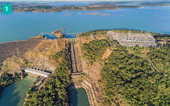 Imagem: Fotografia. Vista aérea de uma barragem, com uma construção do lado direito.  Fim da imagem.