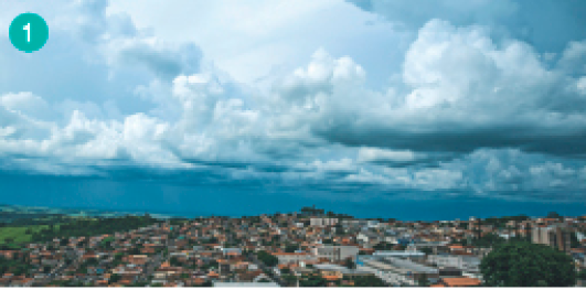 Imagem: Fotografia. Vista aérea de uma cidade. O céu está cinza e coberto por nuvens.  Fim da imagem.
